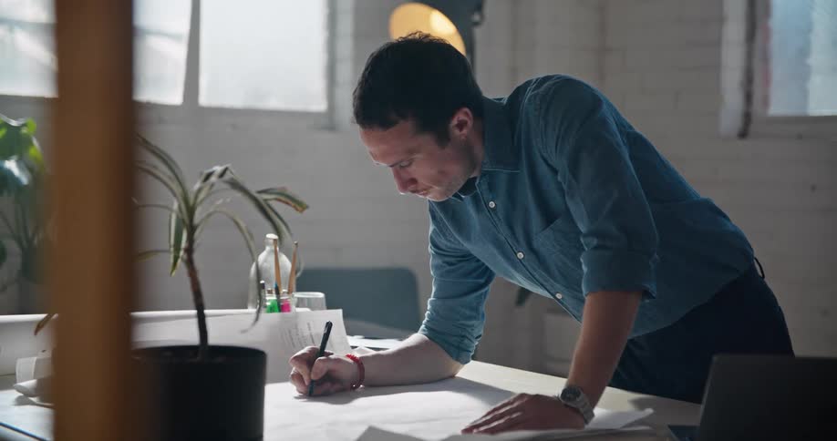 Man in office leans over desk to make notes on paper while consulting laptop
