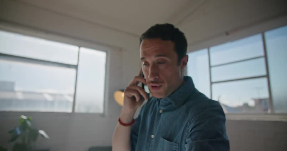A man using a smart phone standing in front of a desk with a wall and windows in the background