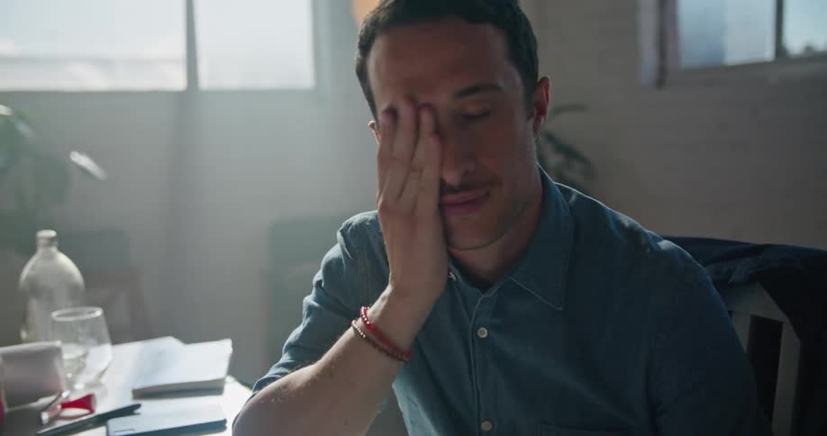 A man is stilling at a desk with a computer in front of him as he turns away and rubs his eyes