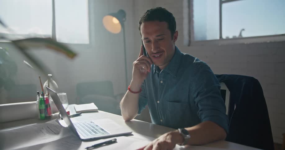 A man is sitting at a desk typing on a computer and talking on the phone