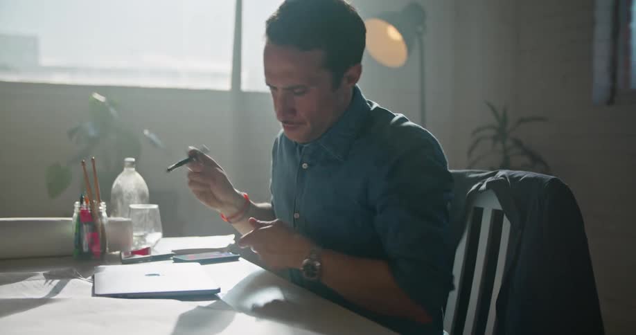 A man sitting at his desk waves to someone on his laptop and then closes it and writes on papers on his desk