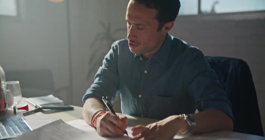 A man in an office sits at a desk and waves to someone on a computer as they video chat and then he talks and writes something down