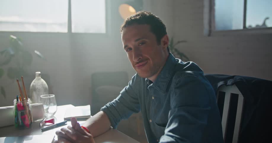 A man smiles into the camera while sitting at a desk indoors during the day