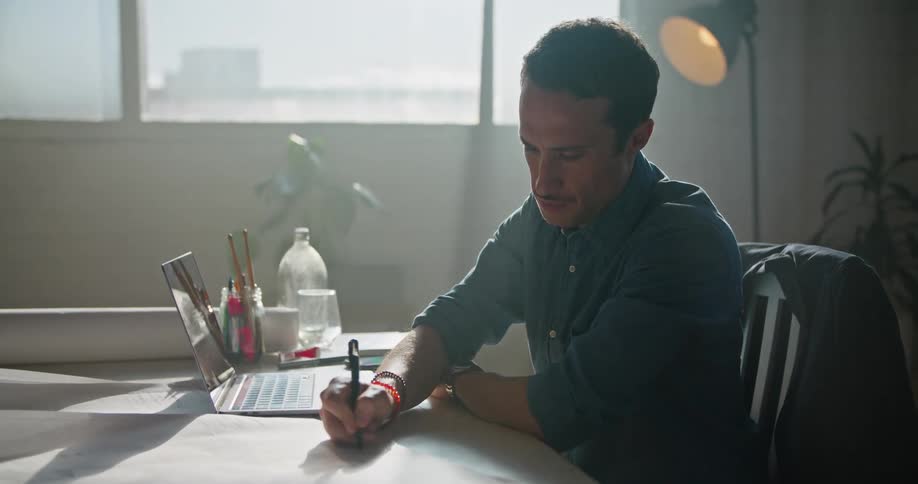 A man in a room sitting on a chair at a desk using a laptop with a wall and window in the background