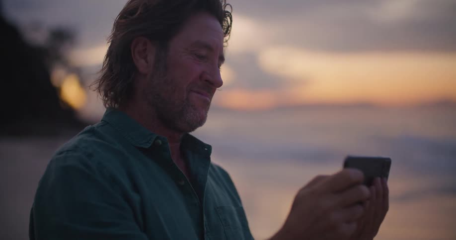 A man is standing on a beach taking a picture with his cell phone