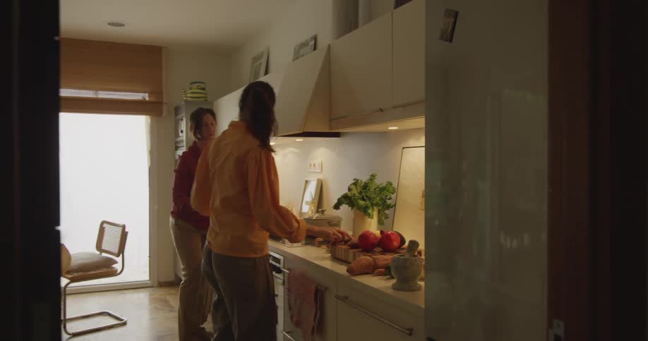 Two women in the kitchen with one cutting vegetables with a wall table and chairs in the background