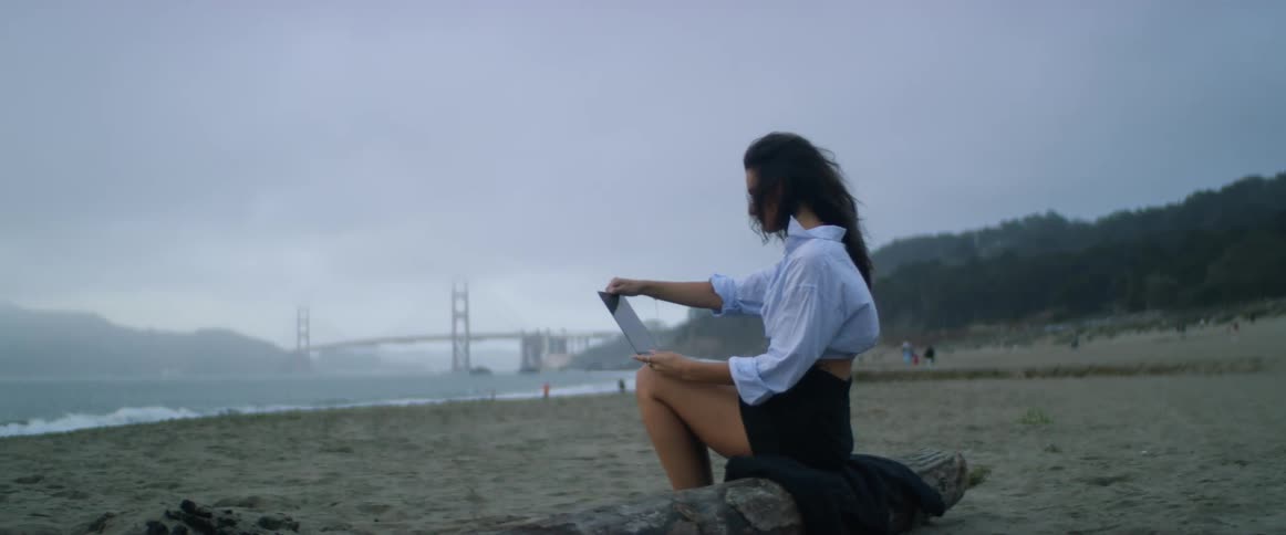 A young, Middle-Eastern woman brushes her long, dark, windblown hair out of her face with her fingers as she sits on the beach working on her laptop with the Golden Gate bridge in the distant fog.
