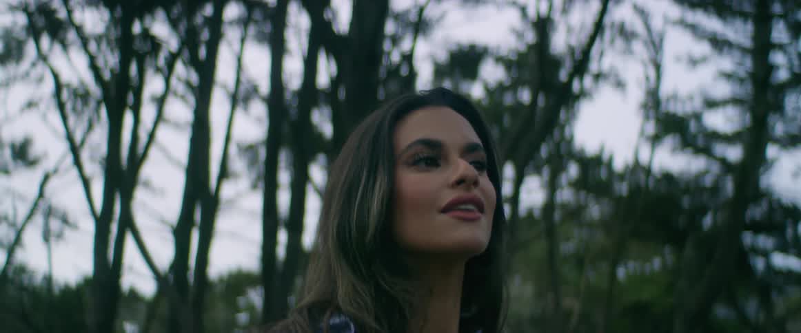 Woman with wavy hair and light blue shirt looks around in the woods smiling