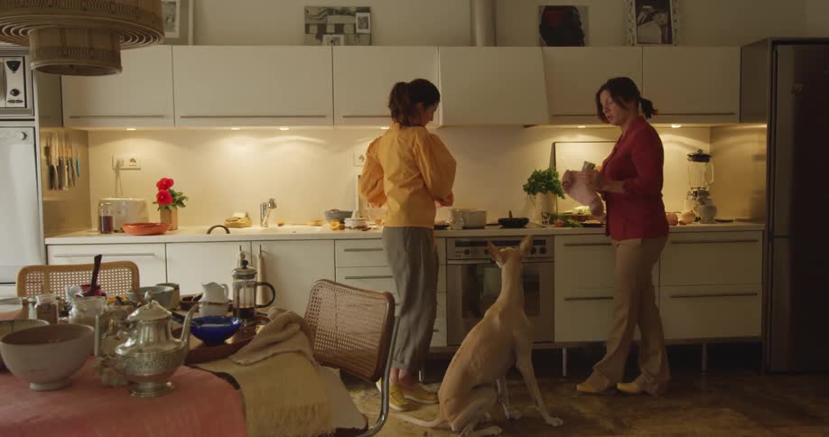 Two women stand in a kitchen with a dog and talk as they prepare food and drink out of mugs