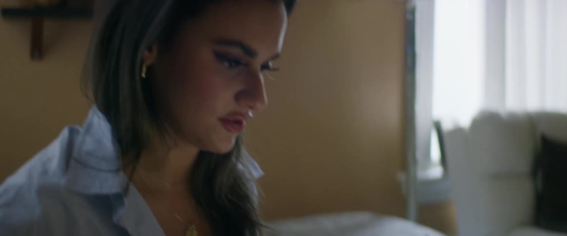 A young, ethnic, business woman sitting on a bed working on a laptop and talking to someone off camera.