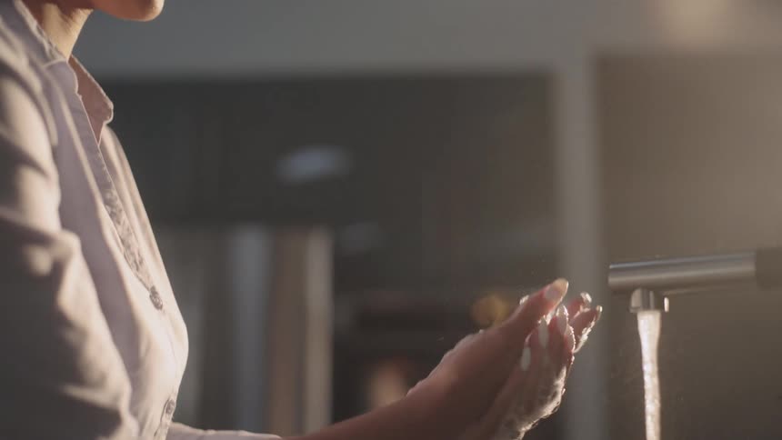 Woman with white shirt and brown wavy hair washes her hands in sink using soap in the kitchen