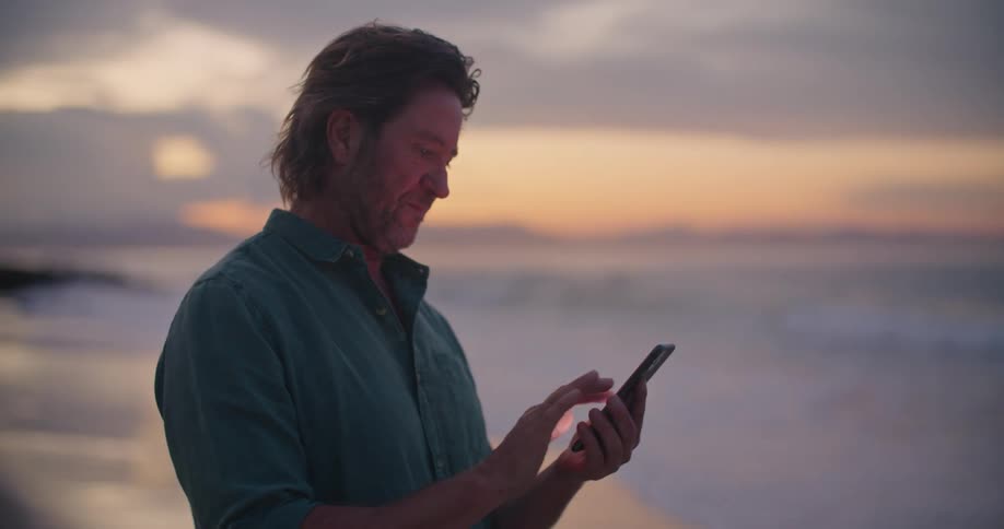 A man is scrolling on a smart phone while standing on an ocean beach after sunset