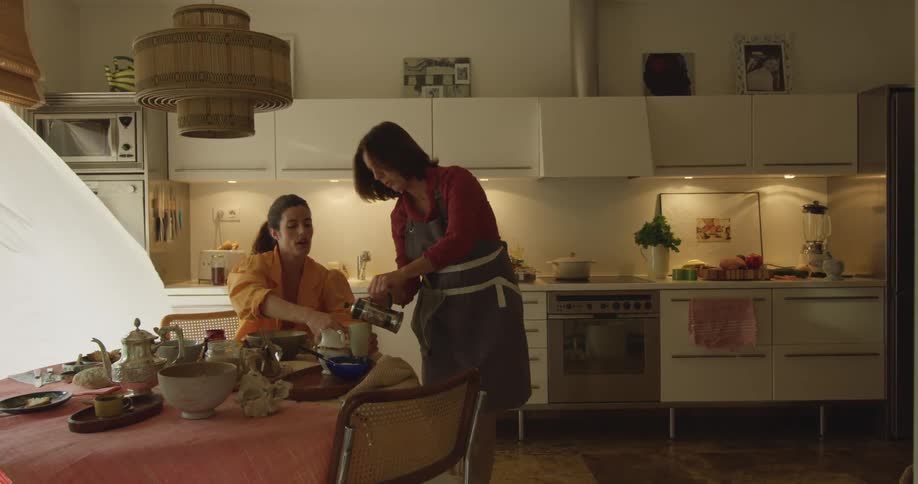 A woman sits at a table in a kitchen as another woman pours her a cup of coffee