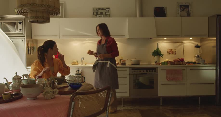 A woman is eating as another walks around the kitchen talking