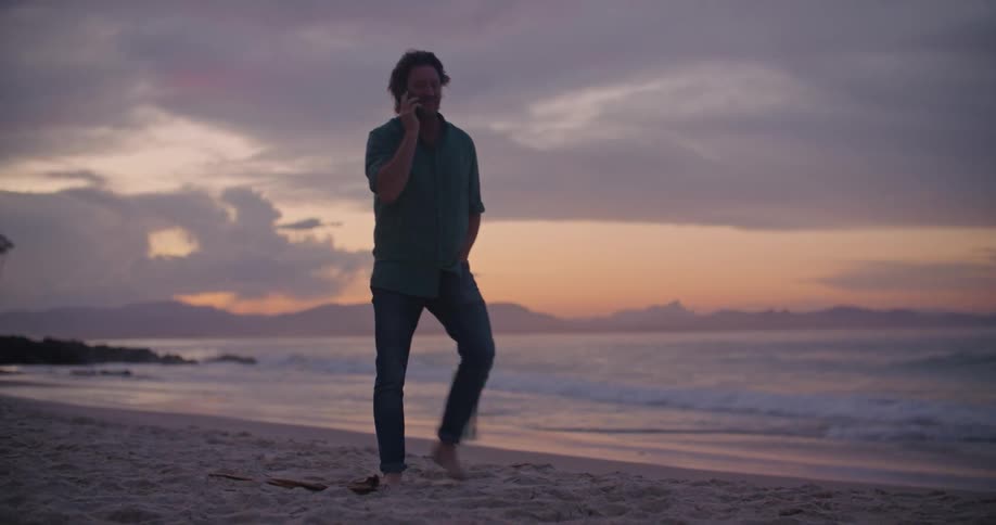 A man talks on his smartphone while walking barefoot along a beach at sunset