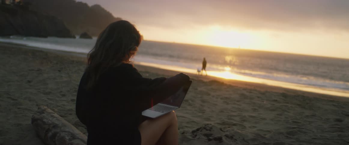 A young, dark-haired woman sitting on the beach at sunset pulls out her laptop as she watches a man and his dog playing in the sand near the waves.