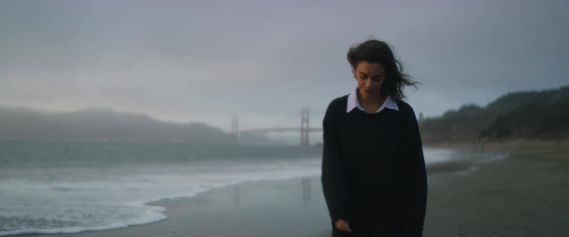 A woman is walking around alone on a windy beach in the evening with waves crashing and the golden gate bridge in the background