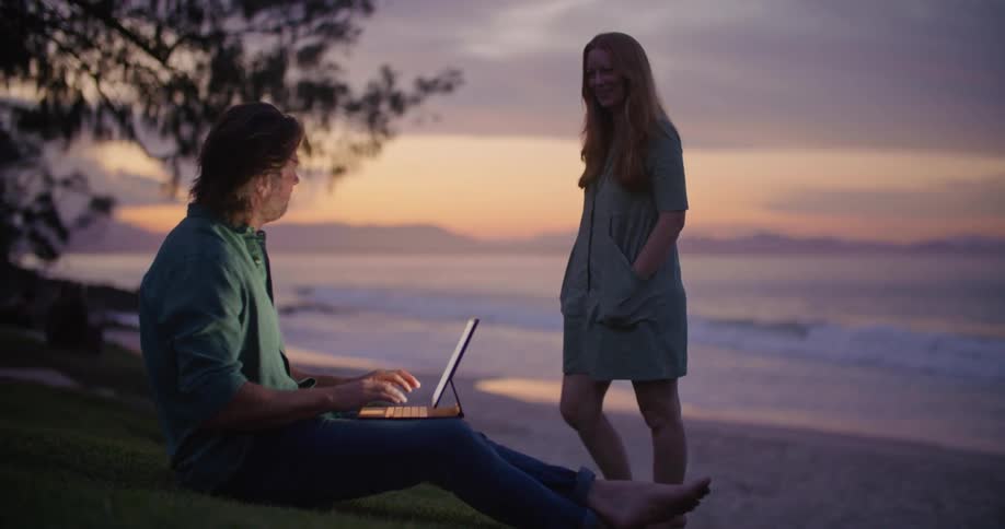 Man with blue shirt and jeans sitting on slope talks to blonde woman with blue dress and uses tablet at the beach next to sea at sunset