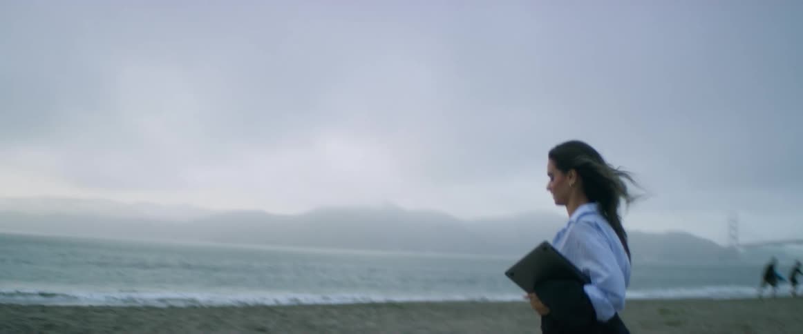 A young, brunette, multicultural woman, in work attire with a laptop in hand, stands on the beach on a foggy day breathing in the fresh air, and then looks at the bridge in the distance as she walks forward to set up a space to sit on the sand.