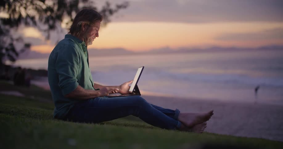 Man with blue shirt and jeans sitting on slope uses tablet at the beach next to sea at sunset