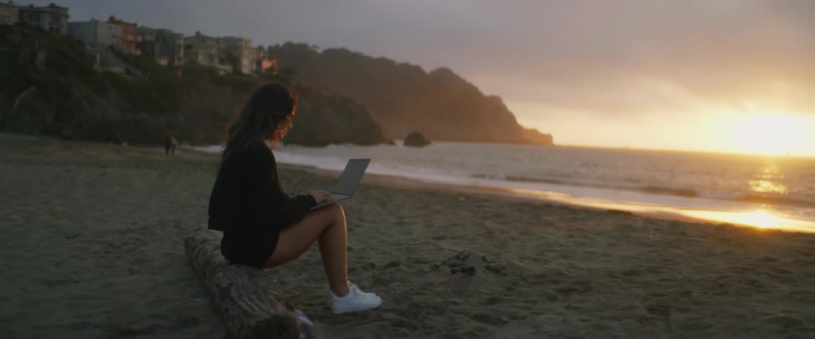 A young, dark-haired woman sits on a log at the beach working on her laptop, with cliffs and buildings in the distance, as the sun sets over the ocean and waves gently hit the shore.