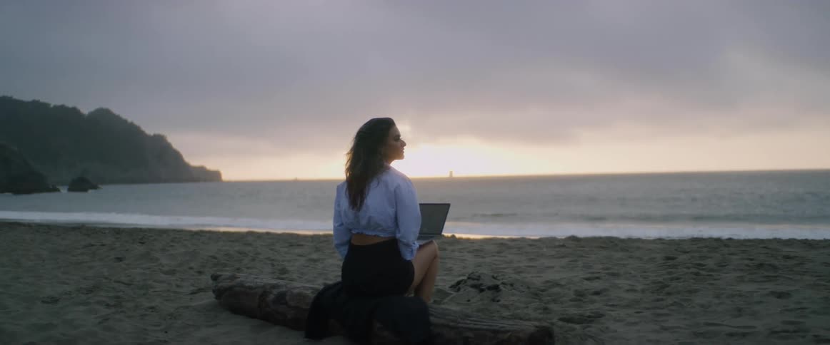 A young woman with dark hair, and her back to the camera, sits on a log on an empty beach with laptop on her lap as she watches the sun rise over the Pacific ocean.