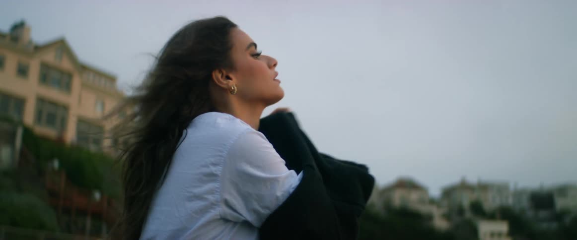 Woman wearing shorts stands from beach sand to put on sweater with hill occupied by buildings and red sky in background