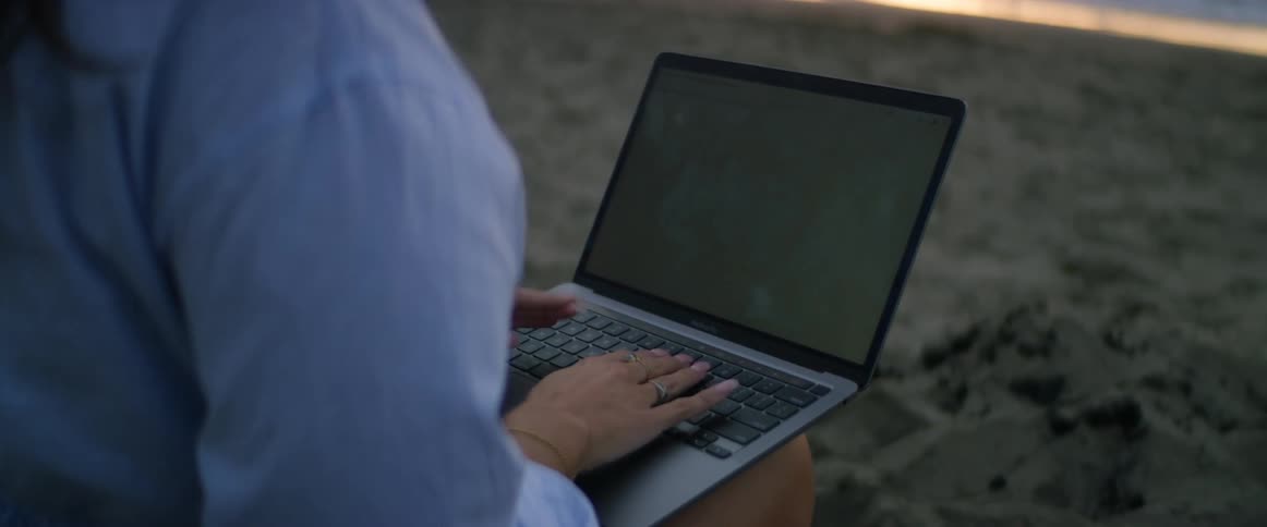 A young, brunette woman with dark hair blowing in the wind is working on her laptop at the beach as she watches the sun set over the Pacific Ocean.