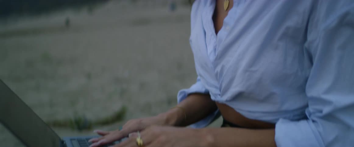 A young, Indian woman with dark hair and multiple earrings types on her laptop as she sits on the beach on a cloudy day with the wind blowing her hair.