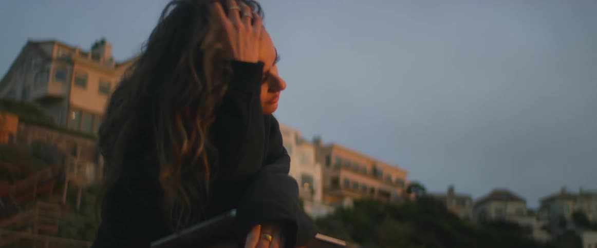 A young, ethnic woman closes her laptop, looks around, and smiles peacefully as she sits outside at sunset with the wind blowing her long, dark hair.
