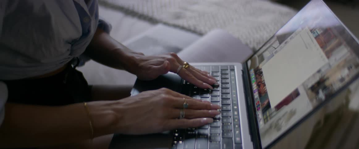A young, multicultural woman with dark hair and eyes sitting on a bed and typing on her laptop while she works from home.