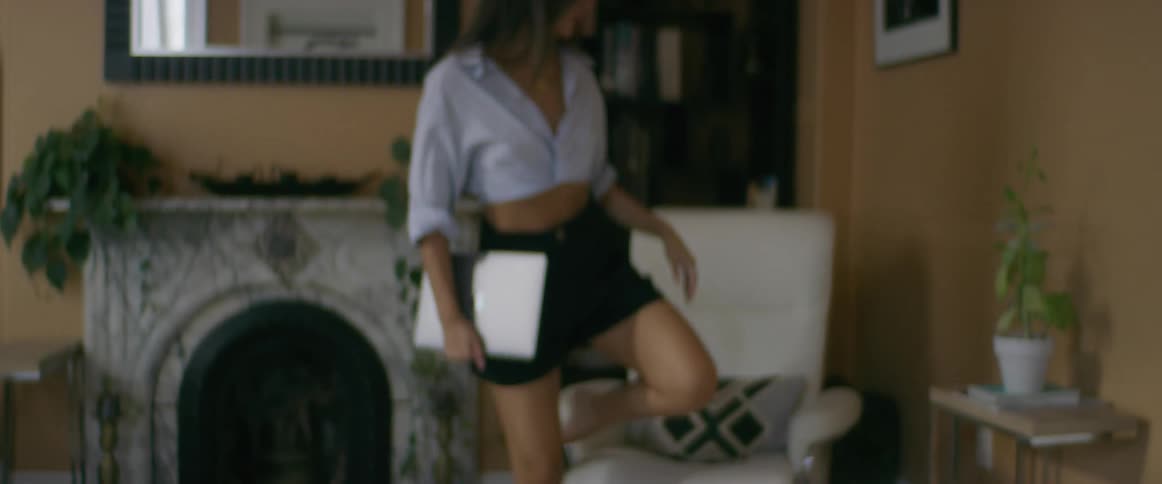 A young, brunette woman stands up from the couch with her computer and moves to an armchair where she can put her bare feet up.