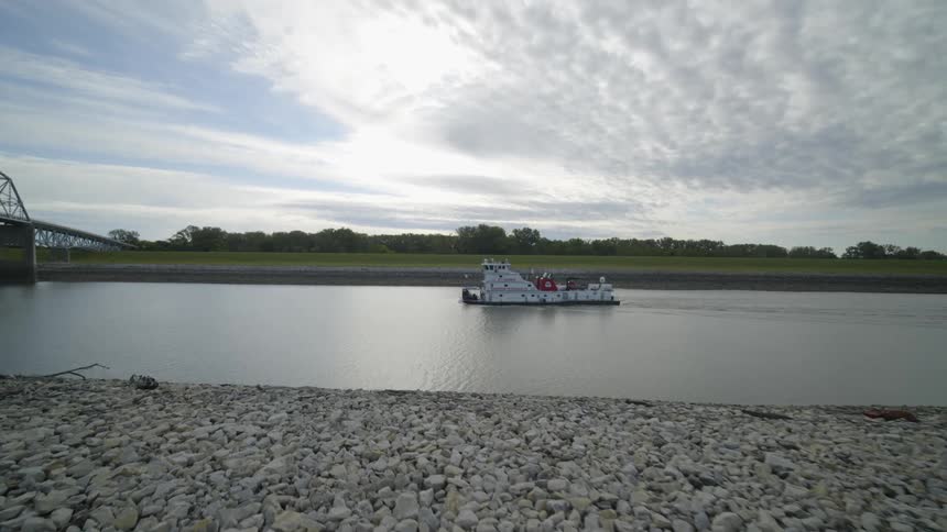 A large pusher tug moves along a calm river toward a bridge