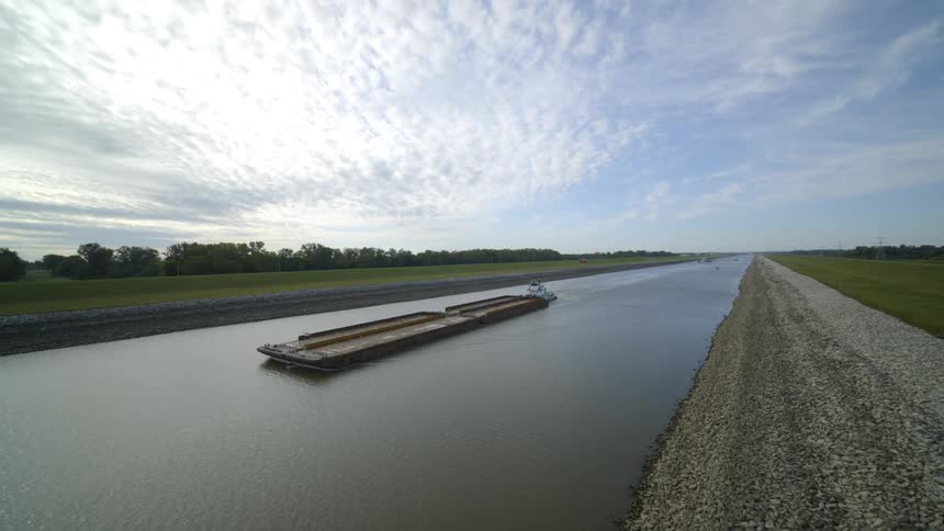 Low approach toward a tug pushing a barge up a water channel surrounded by green fields