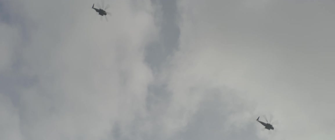 Two helicopters fly in a cloudy sky during the day