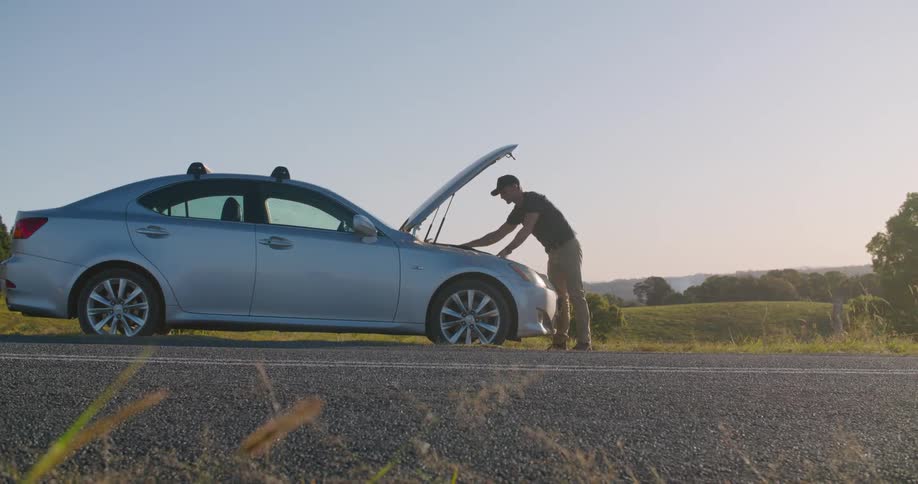 A man with a hat opening the hood of a car with trees the sky and clouds in the background