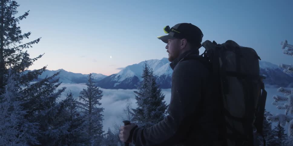 Male mountaineer with cap trek bag and jacket hikes snowy mountain looking around at dusk