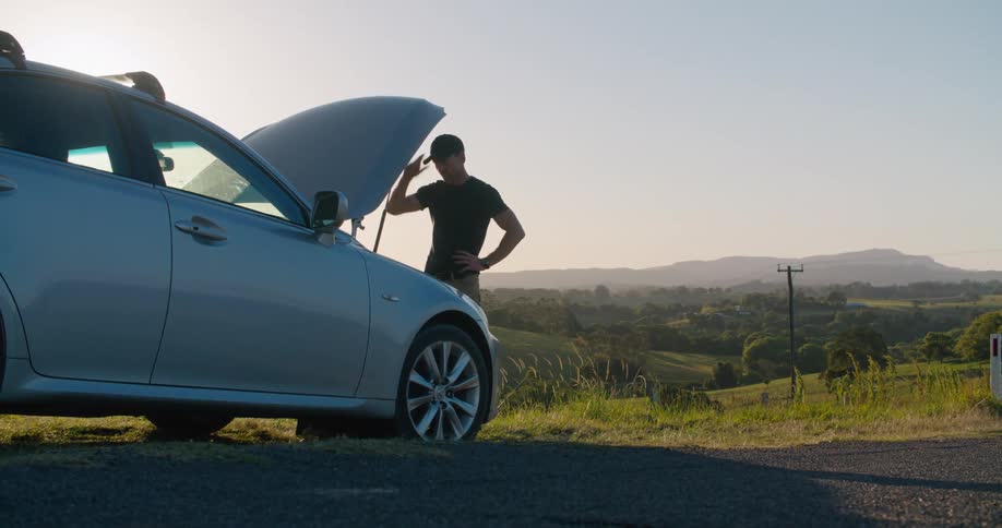 A man checks under the hood of his car thats having problems on the side of a country road in the morning
