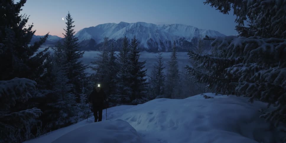 A man wearing a headlight is hiking up a snow covered mountain at night
