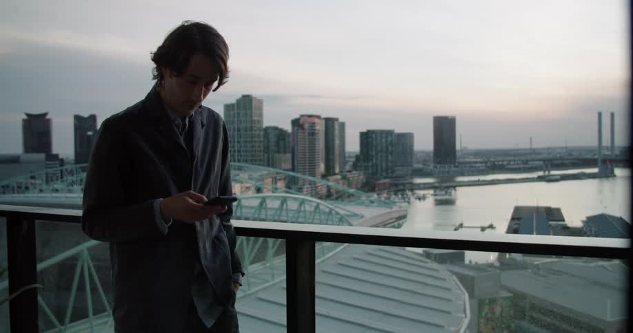 Man stands near rail texting with water and city skyline in background under cloudy red tinted sky
