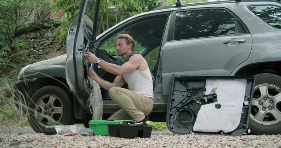 Man crouches working on interior of car door using tool with slope in background