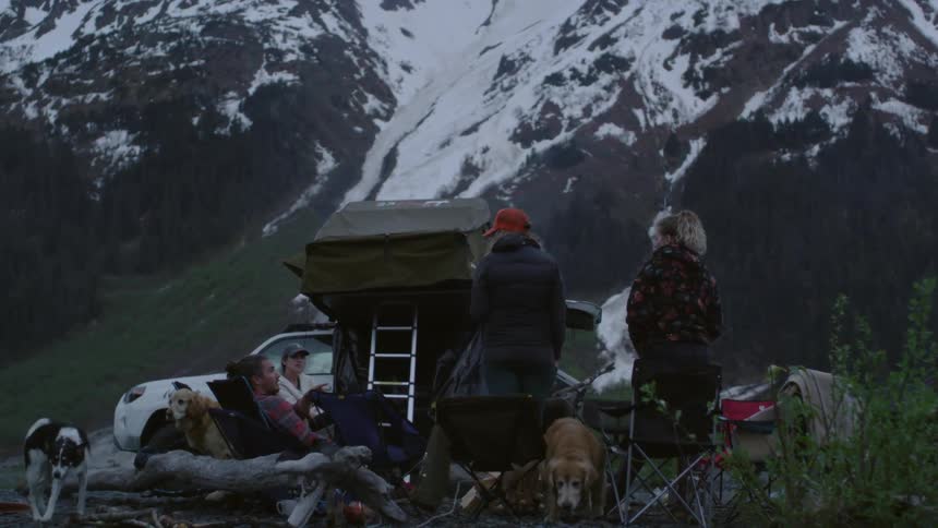 A group of friends sit and stand around a grill as their dogs run around below a mountain