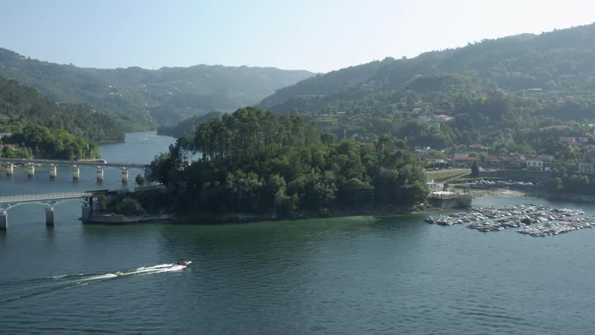 Boats sail across the water as cars travel over a bridge to land with many trees and forested mountains in the distance