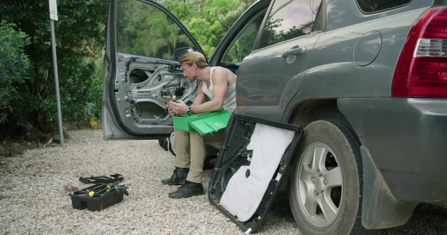 A man looks around and scrolls through his smart phone while sitting in the countryside in his broken down vehicle