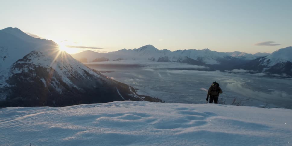 Man hikes up snow covered slope as sun peaks over mountain in mountain range