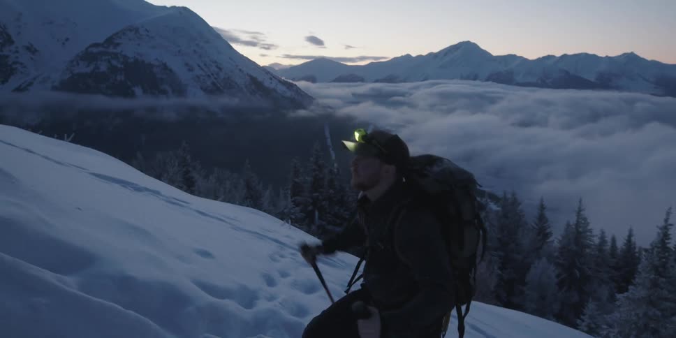 A man with a backpack hiking up a snowy slope with a mountain range the sky and clouds in the background