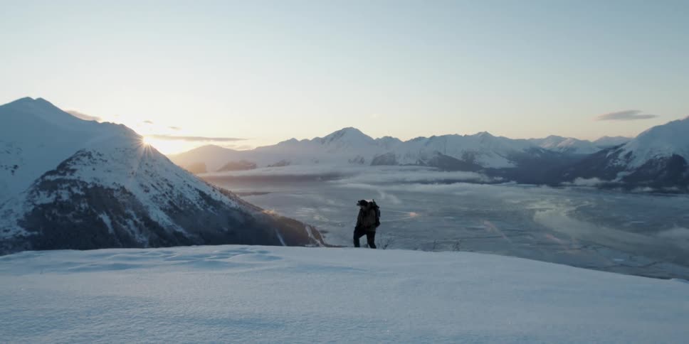 A man is hiking up a snowy mountain with mountains all around with the sun rising in the background