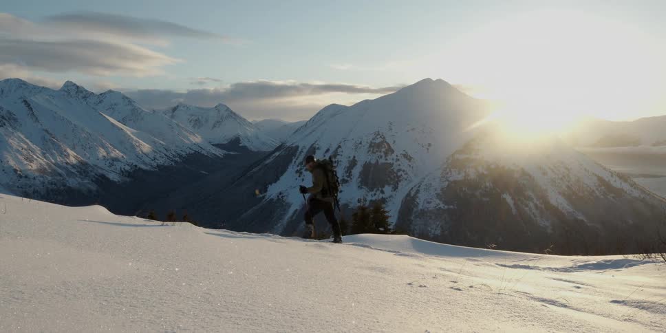 Male mountaineer with trek bag trekking poles hikes snowy mountains while the sun is shining brightly