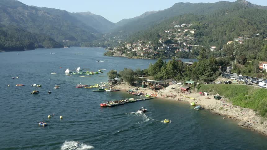 Aerial view of coastal town near the mountains with forest and people on the beach with boats traveling on the sea