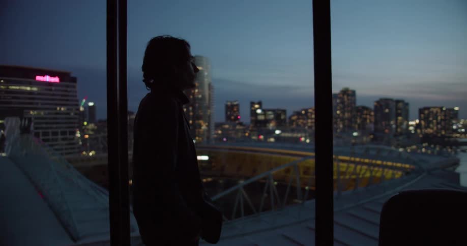 Silhouette of a man standing next to an apartment windown in front of the city skyline at evening